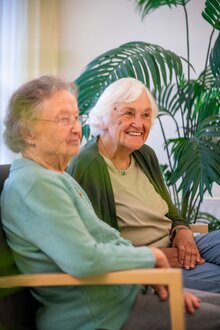 Zwei lächelnde Seniorinnen sitzen zusammen in einer Einrichtung für betreutes Wohnen.