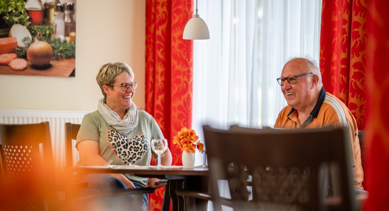 Zwei lachende Senioren sitzen an einem Tisch in einer Wohnanlage.