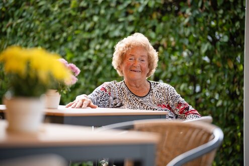 Entspannte, lächelnde ältere Frau sitzt an einem Tisch im Grünen.