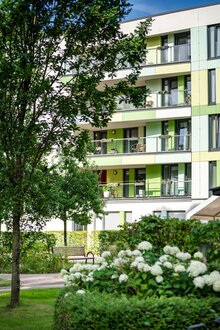 Modernes Wohngebäude einer Wohnanlage mit Balkonen, umgeben von Bäumen, Büschen, weißen Blüten.