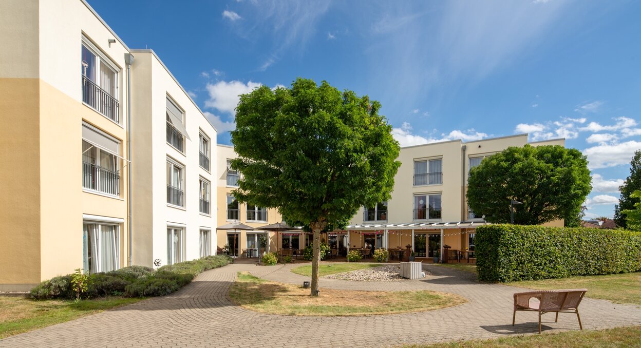 Helle Wohnanlage mit Innenhof, großem Baum und überdachtem Außensitzbereich.
