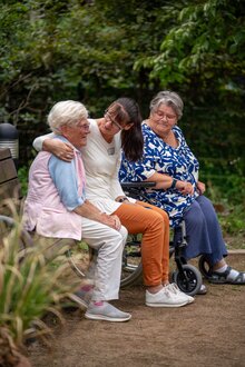 Pflegerin umarmt lächelnd Seniorin auf Bank, weitere Seniorin im Rollstuhl daneben im Grünen.