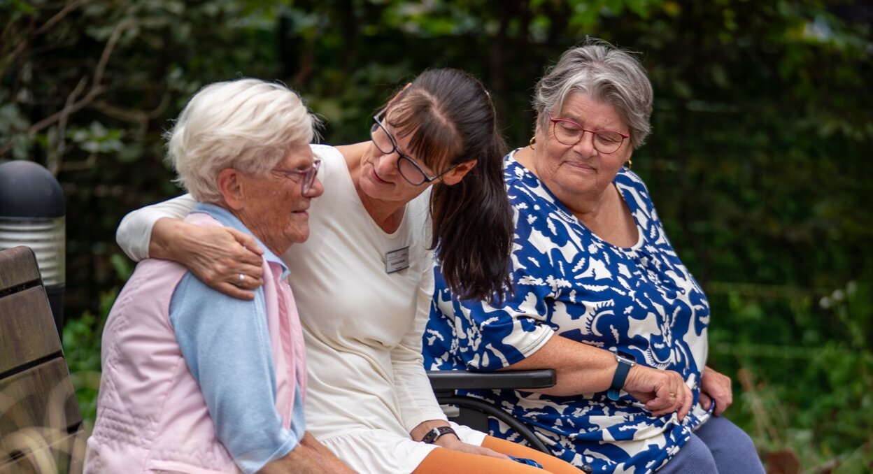 Pflegerin umarmt lächelnd Seniorin auf Bank, weitere Seniorin im Rollstuhl daneben im Grünen.