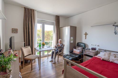 Helles Zimmer einer Wohnanlage mit lesender Seniorin am Fenster und Bett.