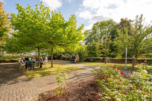 Senioren sitzen in Gruppen unter Bäumen im Garten einer Wohnanlage.