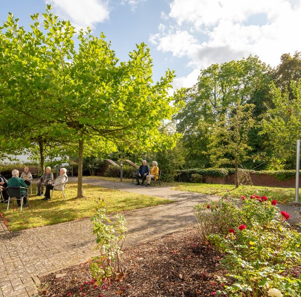 Senioren sitzen in Gruppen unter Bäumen im Garten einer Wohnanlage.