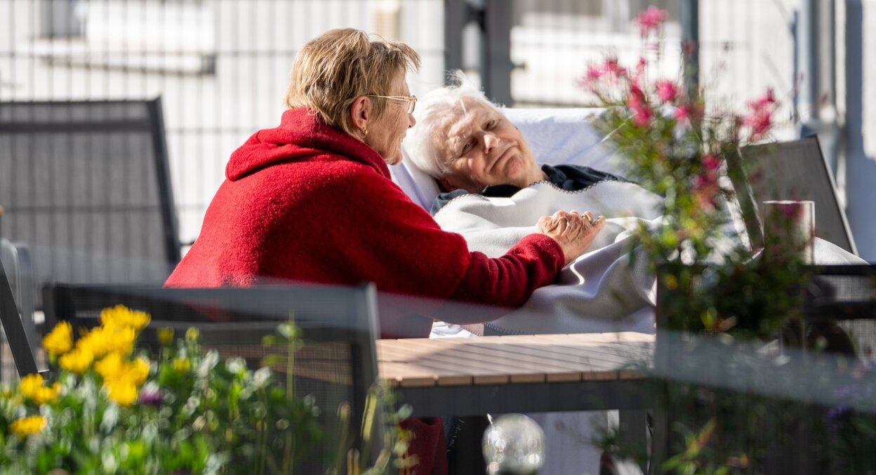Frau hält Hand einer liegenden Seniorin im Freien.