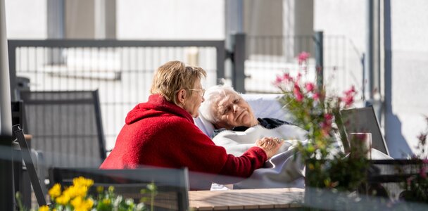 Frau hält Hand einer liegenden Seniorin im Freien.