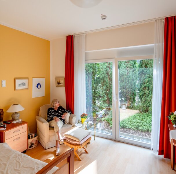 Ältere Frau entspannt in hellem Wohnraum mit Gartenblick bei betreutem Wohnen.