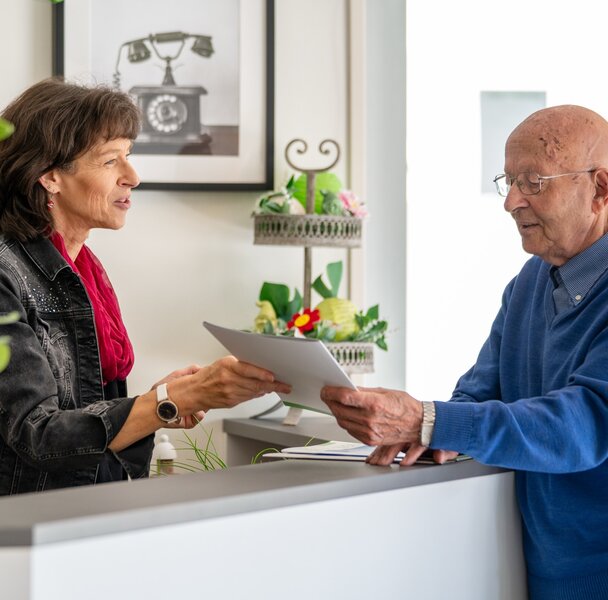 Frau übergibt älterem Mann Unterlagen am Empfang einer betreuten Wohnanlage.