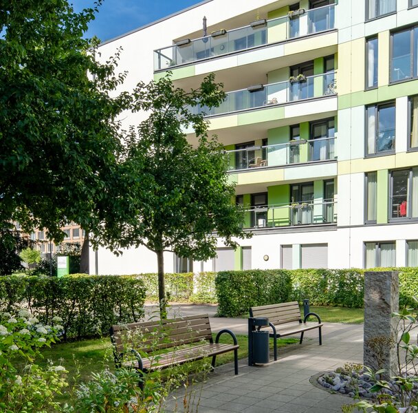 Modernes integra Wohngebäude mit grün-gelber Fassade, Balkonen, Grünflächen und Bänken.