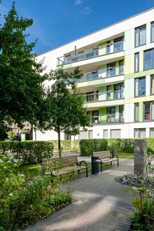 Modernes integra Wohngebäude mit grün-gelber Fassade, Balkonen, Grünflächen und Bänken.