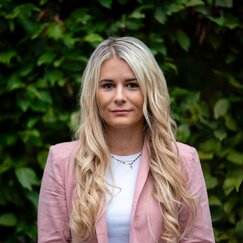 Mitarbeiterin mit blonden Haaren im rosa Blazer vor grüner Hecke.