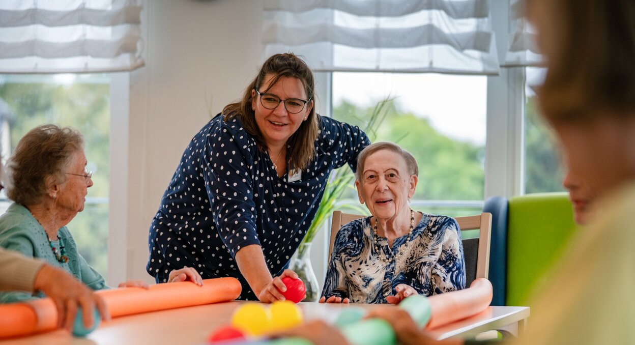 Lachende Betreuerin macht eine Übung mit Senioren am Tisch in einer Wohnanlage.