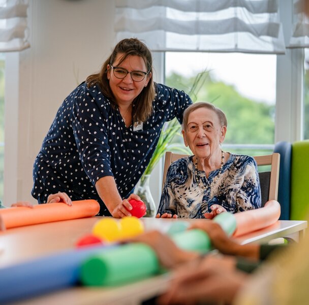 Lachende Betreuerin macht eine Übung mit Senioren am Tisch in einer Wohnanlage.