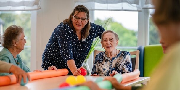 Lachende Betreuerin macht eine Übung mit Senioren am Tisch in einer Wohnanlage.