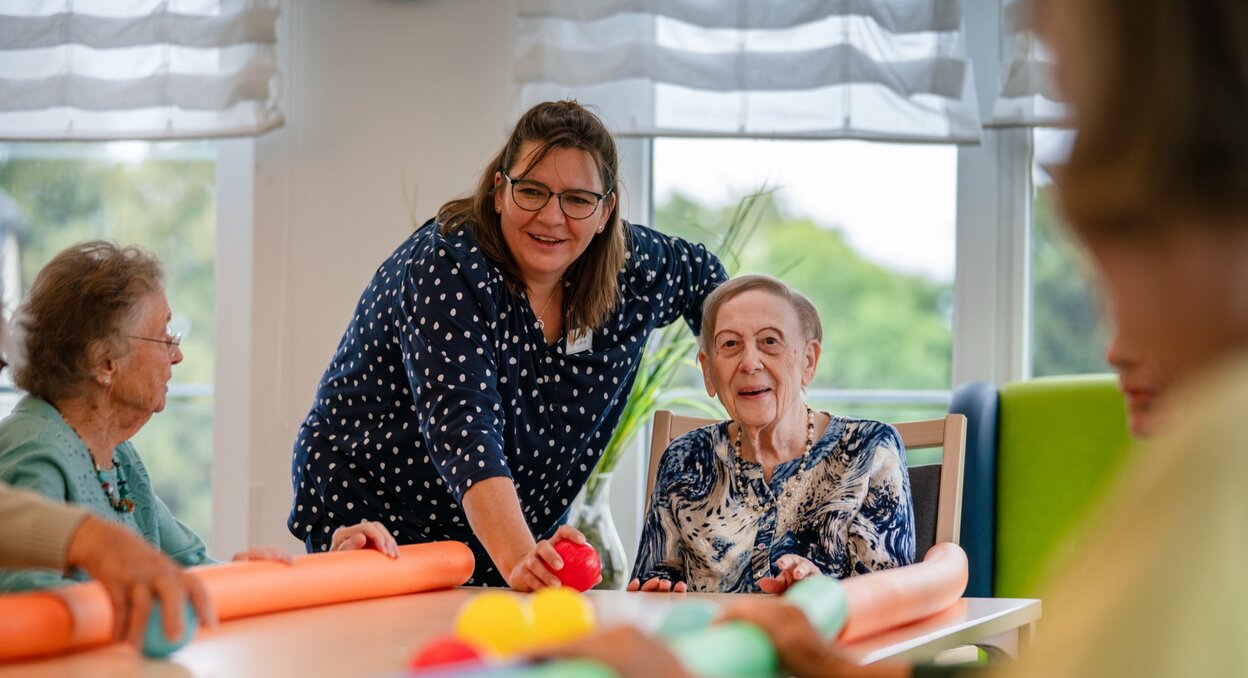 Lachende Betreuerin macht eine Übung mit Senioren am Tisch in einer Wohnanlage.
