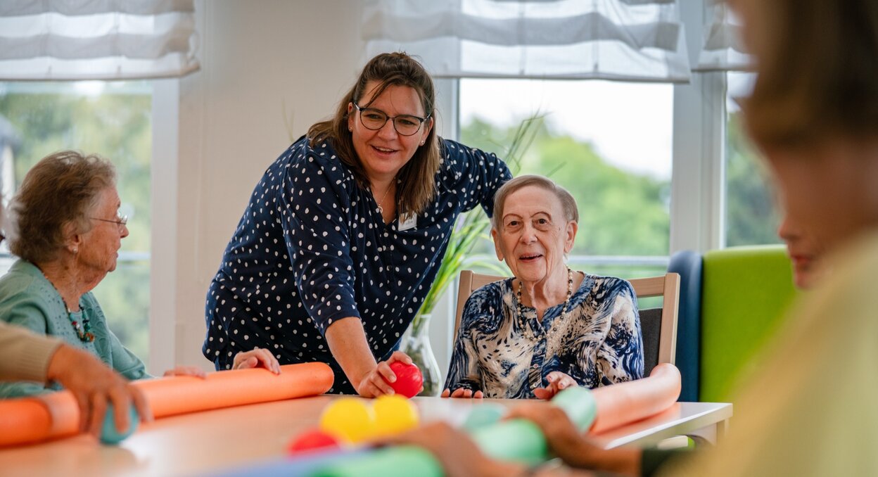 Lachende Betreuerin macht eine Übung mit Senioren am Tisch in einer Wohnanlage.
