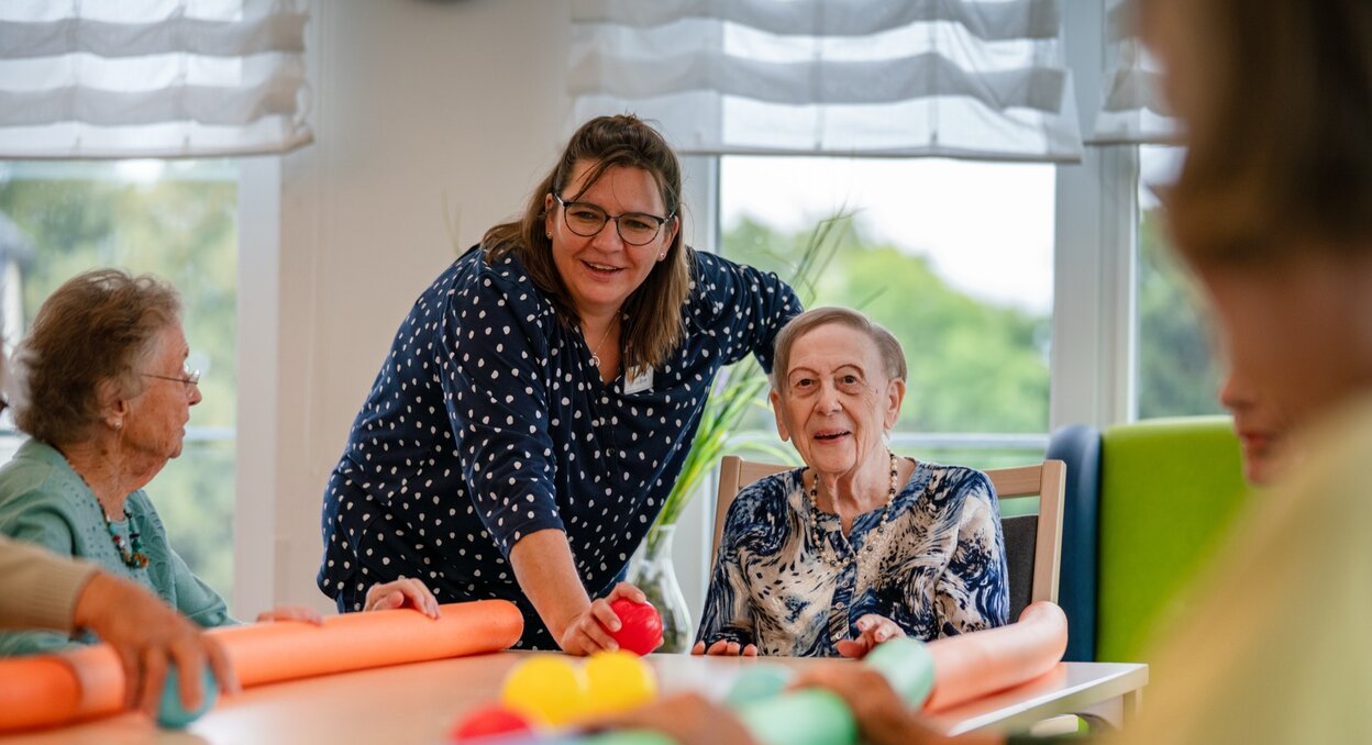Lachende Betreuerin macht eine Übung mit Senioren am Tisch in einer Wohnanlage.