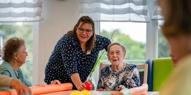 Lachende Betreuerin macht eine Übung mit Senioren am Tisch in einer Wohnanlage.