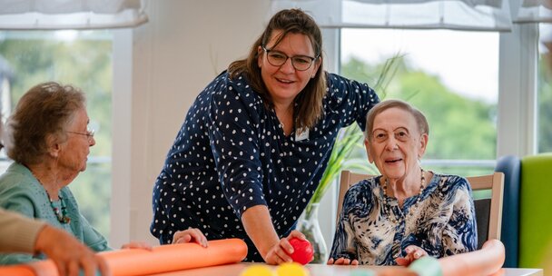 Lachende Betreuerin macht eine Übung mit Senioren am Tisch in einer Wohnanlage.