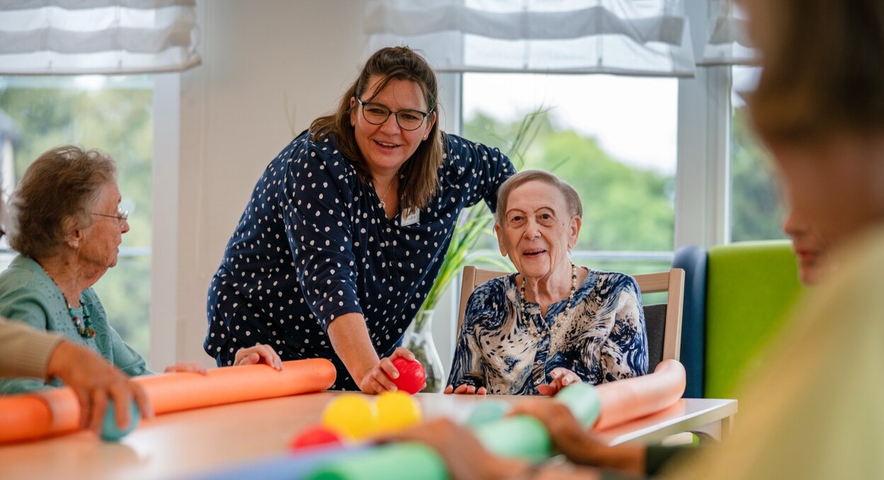 Lachende Betreuerin macht eine Übung mit Senioren am Tisch in einer Wohnanlage.