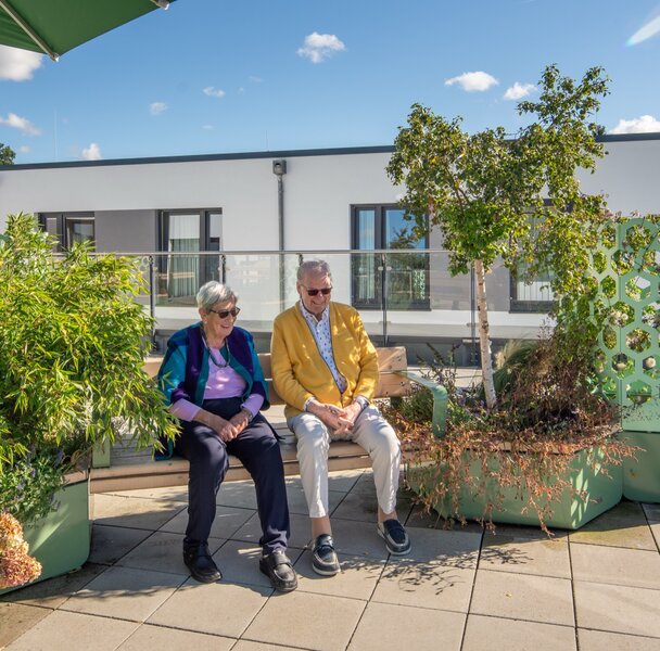 Zwei Senioren sitzen auf einer Bank auf einer grünen Dachterrasse einer Wohnanlage.