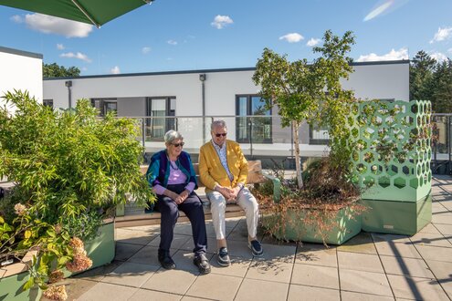 Zwei Senioren sitzen auf einer Bank auf einer grünen Dachterrasse einer Wohnanlage.